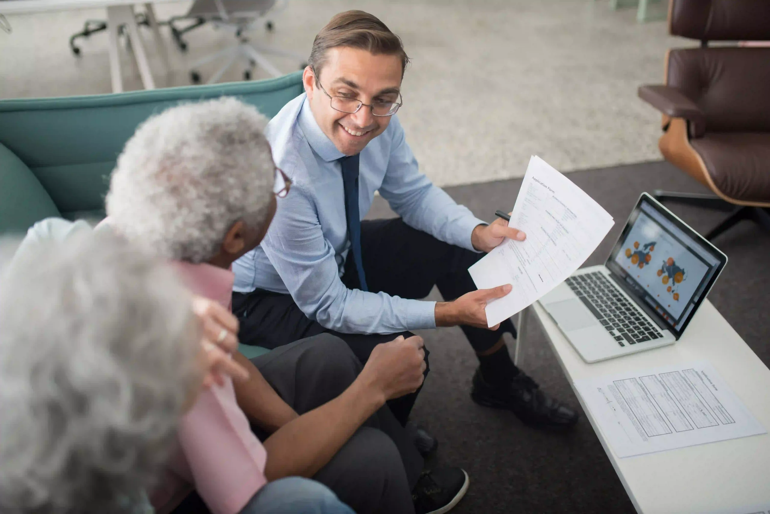 Smiling financial advisor showing an application form and laptop data to a senior couple during a consultation.